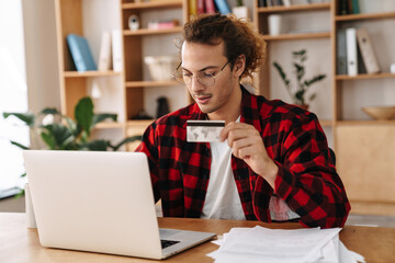 Handsome focused guy holding credit card and working with laptop