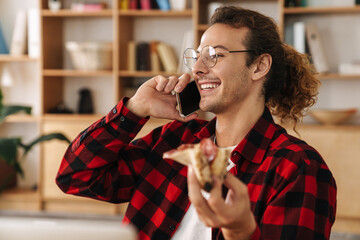 Handsome joyful guy talking on cellphone while eating pizza at office