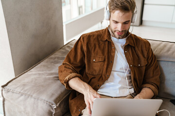 Fototapeta premium Handsome focused guy using headphones and laptop while sitting on sofa