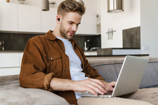 Handsome focused guy using earphones and laptop while sitting on sofa