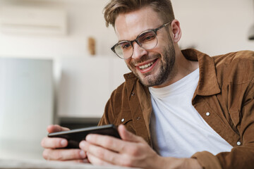 Smiling guy playing online game on cellphone while sitting on sofa