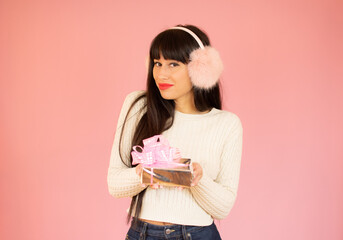 Beautiful young woman in warm sweater and earmuffs holding a gift box on color background