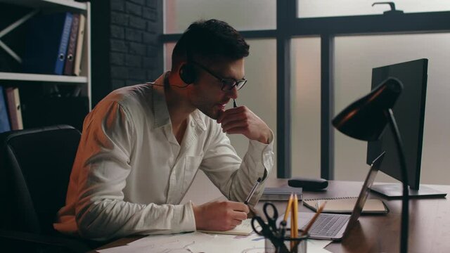Serious businessman in headset making online conference call, talking, reading information on laptop, taking notes. Corporate sales agent advertising services to clients, giving technical support.