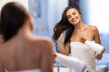 Young woman holding hair brush and drying hair at mirror in bathroom
