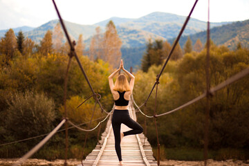 Young woman standing in tree yoga pose, meditating on bridge over mountain river.