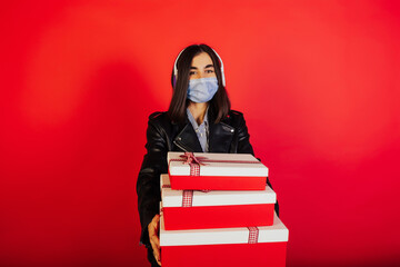 Girl with headphones wear medical mask and hold gift boxes on red background. She holding big three boxes with bows. Christmas gifts. Trendy style. Copy space.