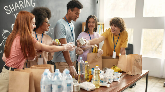 Happy young volunteers group in gloves collecting, sorting food in paper bags, Diverse team working together on donation project in charitable organization office