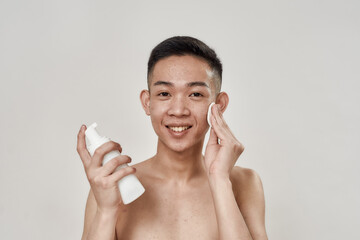 Portrait of shirtless young asian man with problematic skin cleaning his face with lotion using...