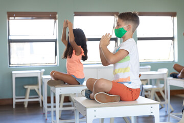 Boy and girl wearing face mask practicing yoga while sitting on the desk in class