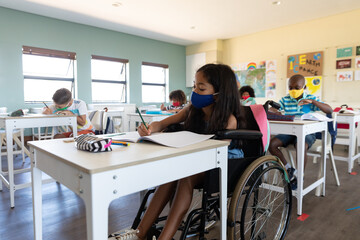 Disable girl wearing face mask writing while sitting on wheelchair in class