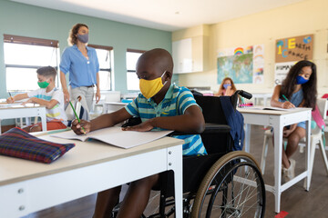 Disable boy wearing face mask writing while sitting on his wheelchair in class