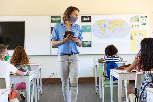 Female teacher wearing face mask holding digital tablet teaching to students in class