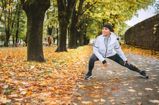 Fit athletic woman doing workout before jogging in the autumnal city park on the kids playground. Young fitness female runner stretching legs while warming up. Active runners people concept image.