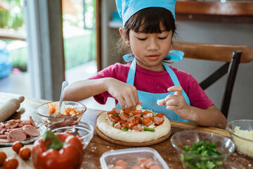 Child in Asian chief hat makes pizza with tomatoes and grated cheese - cooking, food, and pleasure concept