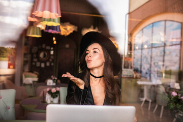 Attractive woman with long curly hair in black hat sitting in cafe with laptop.