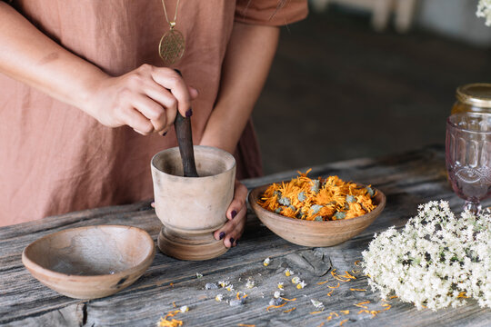 Midsection of woman crushing flowers and herbs in mortar with pestle on wooden table
