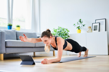 Woman extending arm during plank pose while learning exercise on internet through tablet PC