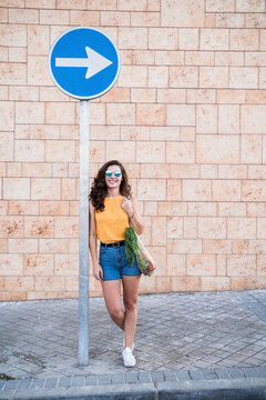 Smiling young woman leaning on directional sign against wall in city