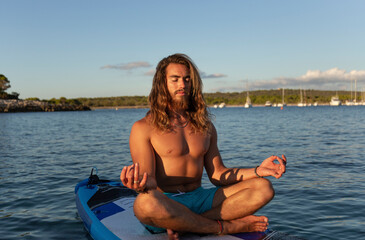 Young man with long hair meditating while sitting on paddleboard at sea against sky during sunset
