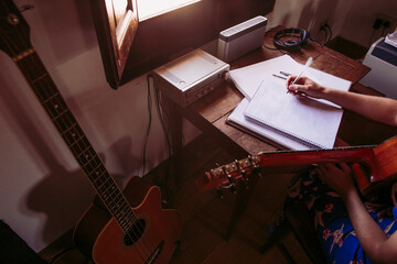 Young woman writing in book while practicing guitar in living room