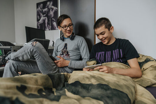 Teenage boys sitting on bed, having fun using portable devices