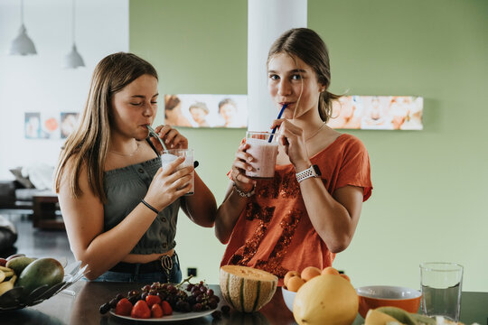 Teenage girls standing in kitchen drinking fresh fruit smoothies with drinking straws