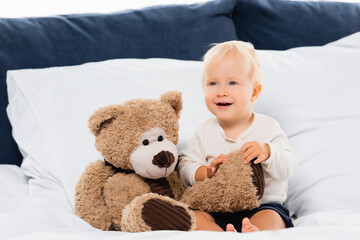 Selective focus of toddler boy smiling near teddy bear on bed on white background