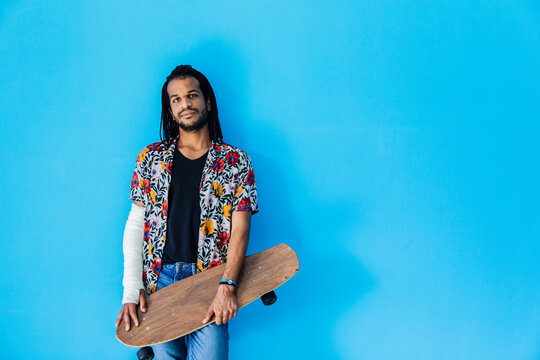 Mid adult man with fractured arm holding skateboard against blue background