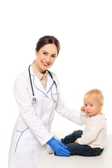 Smiling pediatrician looking at camera near toddler on table isolated on white