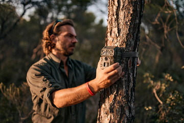 Mature man holding trail camera on tree trunk while standing in forest