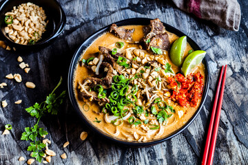 Bowl of red curry soup with rice noodles, beef, vegetables, scallion, peanuts and lime