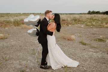 Smiling groom with heart shape balloons kissing bride against sky