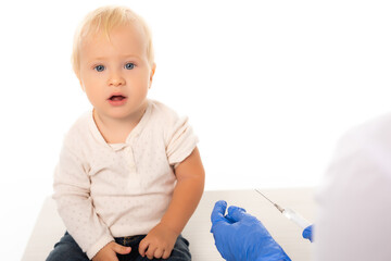 Selective focus of toddler boy looking at camera near doctor with syringe isolated on white