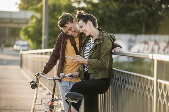 Smiling boyfriend and girlfriend looking at smart phone while standing with bicycle in city