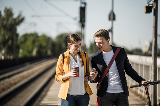Young man showing smart phone to female friend while walking on railroad station platform