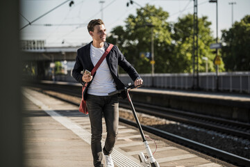 Smiling young man looking away while walking with push scooter on railroad station platform