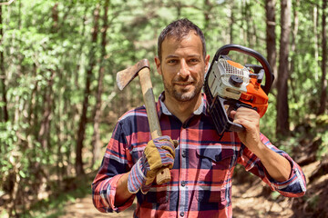 Smiling lumberjack standing with axe and chainsaw in forest
