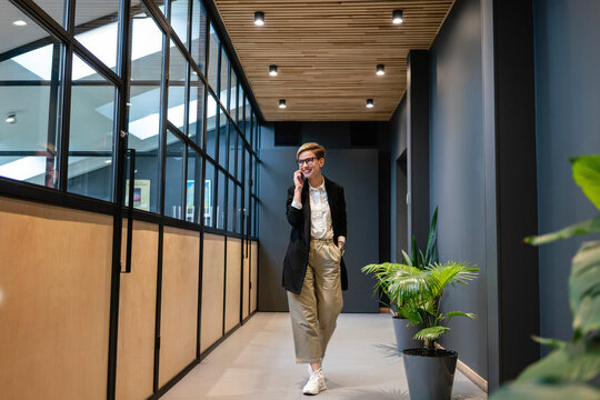 Smiling beautiful businesswoman talking on smart phone while walking in illuminated corridor at office