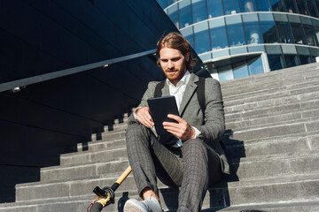 Young male entrepreneur using digital tablet while sitting with electric push scooter on steps in financial district