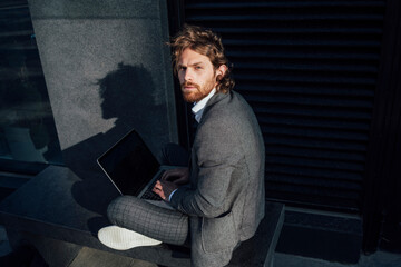Confident young businessman with laptop sitting on bench at financial district in city