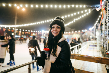 Pretty positive lady stands on the background of the rink with a cup of warming drink and sparklers...