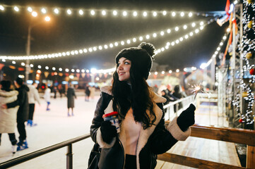 A happy girl in warm clothes stands by the rink with cups of hot drink in her hand, looks away,...