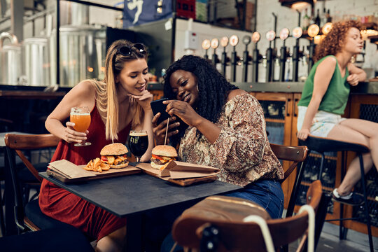 Happy female friends with smartphone having burgers in a pub