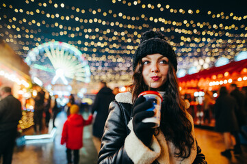 A pretty girl in a hat and winter clothes walks down the Christmas street in the evening and drinks a hot drink from a paper cup. Lady stands at a decorated Christmas market in the evening.