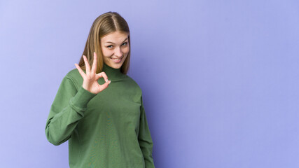 Young blonde woman isolated on purple background winks an eye and holds an okay gesture with hand.