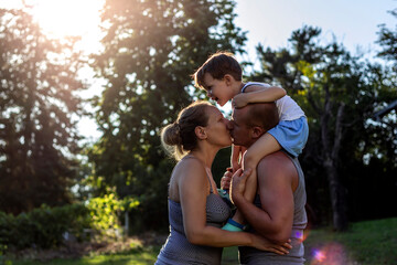 Photo of a happy family enjoying time together. Young loving father carrying on shoulders smiling son, kissing his wife while spending free time in the nature. A young family with a toddler having fun