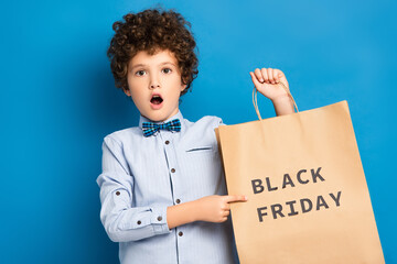 shocked and curly boy pointing with finger at shopping bag with black friday lettering on blue