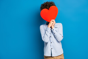 curly boy covering face while holding red paper cut heart on blue