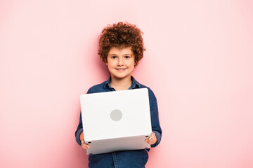 joyful and curly boy holding laptop on pink