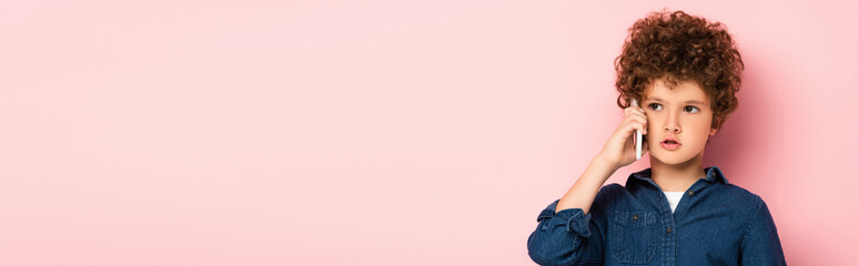 horizontal image of curly boy in denim shirt talking on mobile phone and looking away on pink